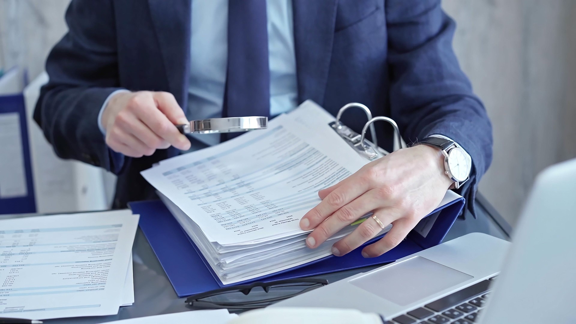 Adult businessman or auditor examining documents with magnifying glass. Professional in a suit scrutinizes financial reports, detail-focused work concept